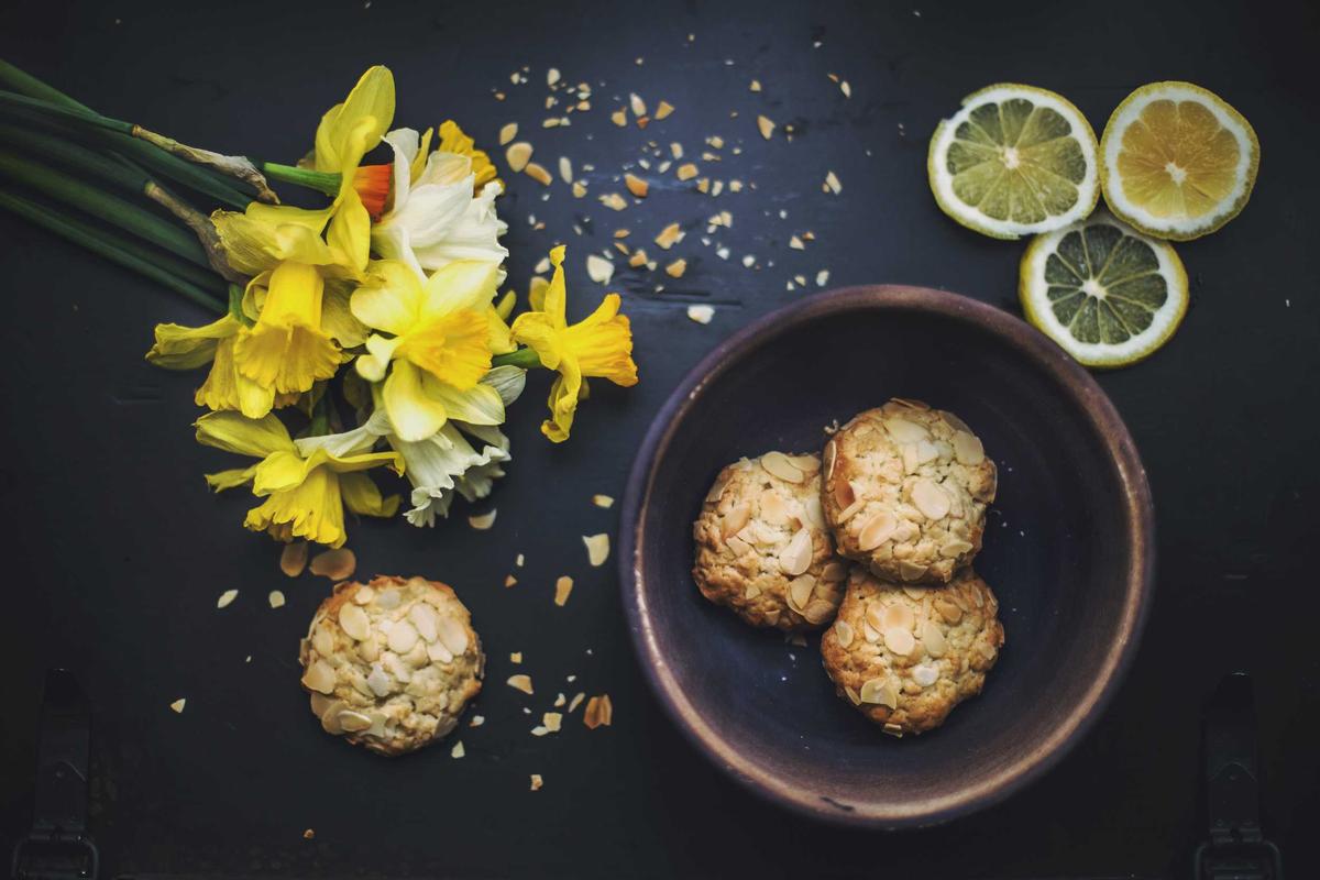 Des jonquilles jaune vif, des tranches de citron et des biscuits aux amandes maison sur un fond sombre.