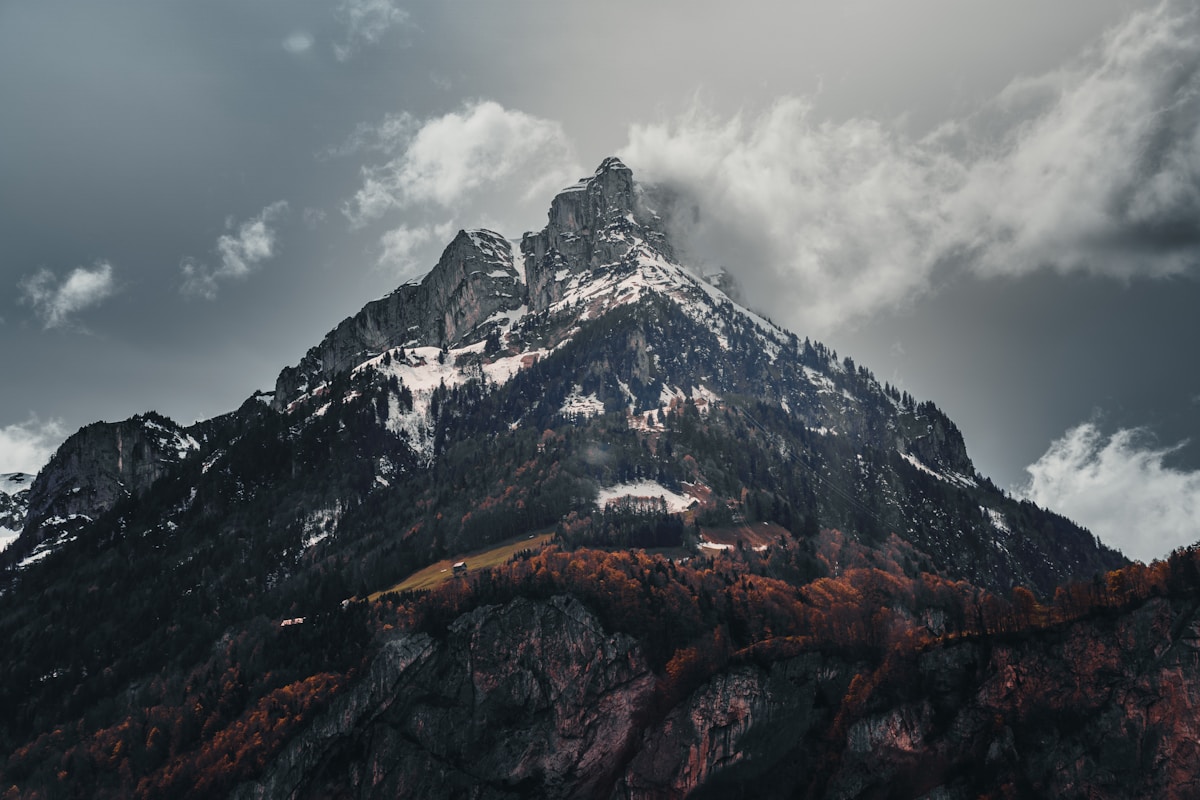 Montagne enneigée entourée de forêts automnales sous un ciel nuageux.