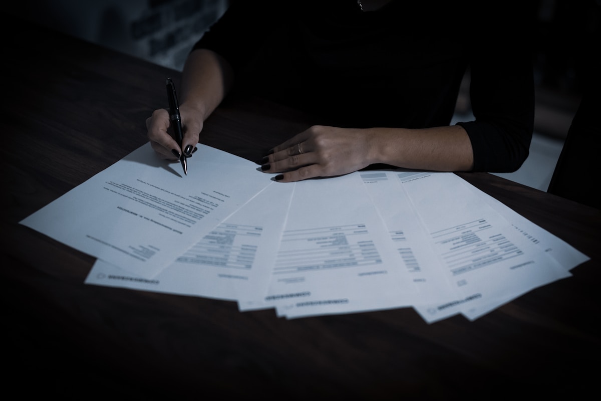 Mains tenant un stylo sur des documents éparpillés sur un bureau sombre.